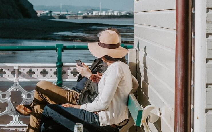 A woman wearing a large hat sitting next to her friend at bench enjoying the water nearby