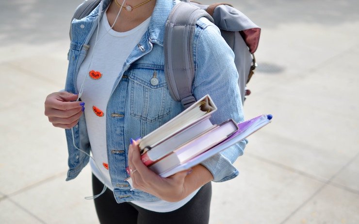 Girl wearing jean jacket while holding books, backpack and listening to music on headphones