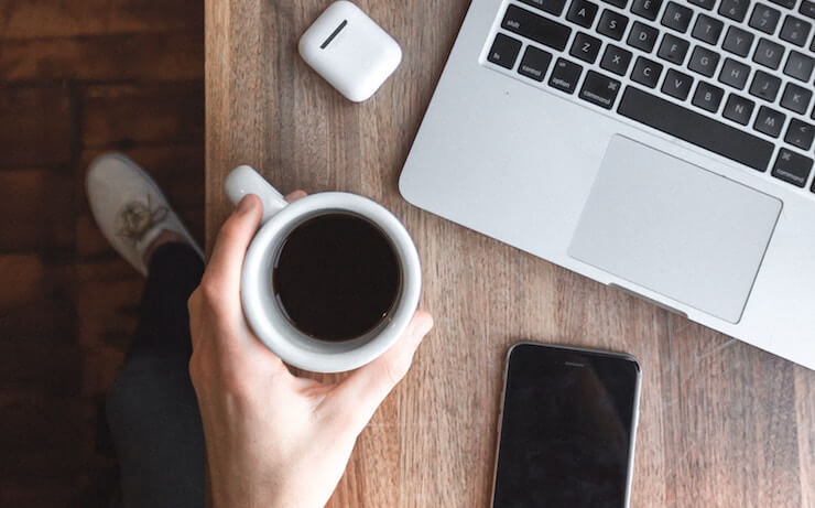 Person relaxing holding a cup of coffee and using their phone and laptop on a table